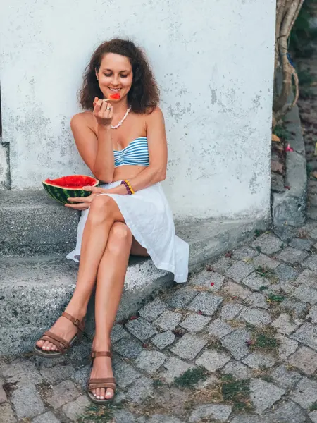 Dame med hvitt skjørt og bikinitopp sitter på steintrapp og spiser vannmelon 