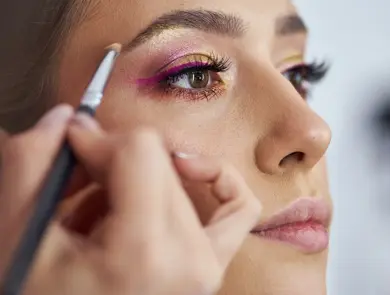 Dedicated young female make up artist applying a purple eyeliner under customer's eye, while giving her professional service at her make up studio