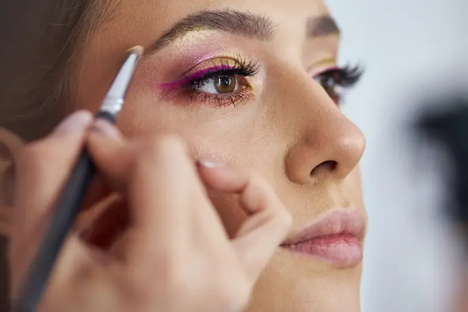 Dedicated young female make up artist applying a purple eyeliner under customer's eye, while giving her professional service at her make up studio