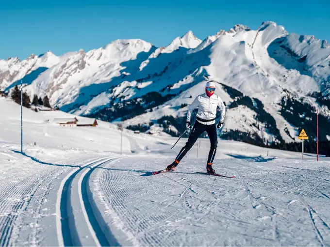 Langrennsløype og mann som går på langrenn, snødekte fjell i bakgrunnen, blå himmel og sol.