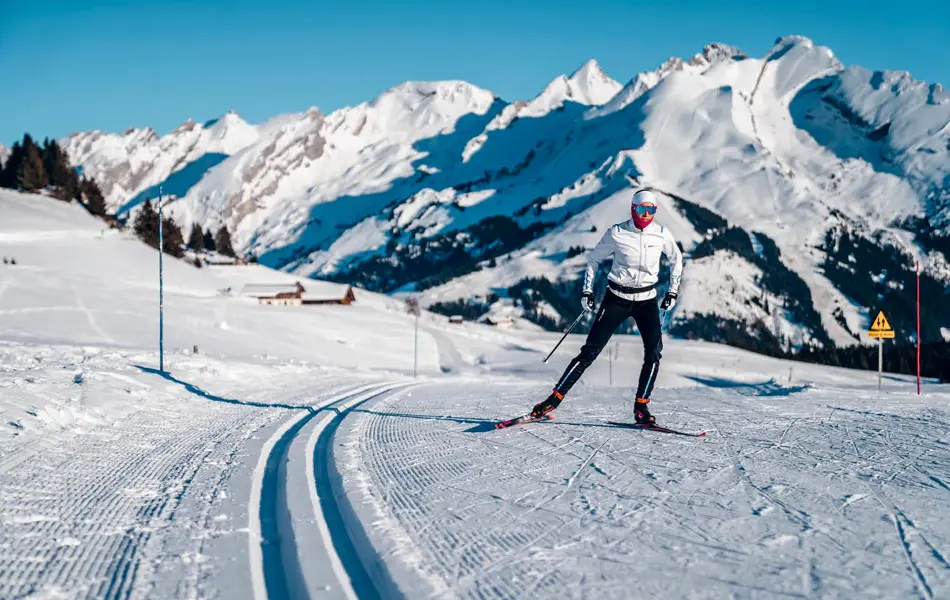 Langrennsløype og mann som går på langrenn, snødekte fjell i bakgrunnen, blå himmel og sol.