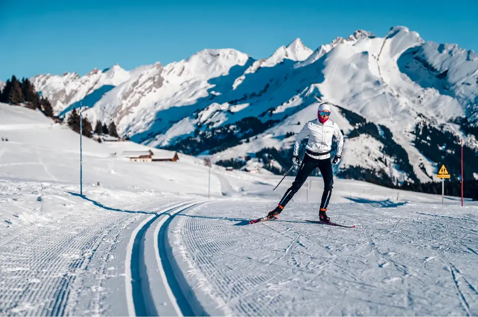 Langrennsløype og mann som går på langrenn, snødekte fjell i bakgrunnen, blå himmel og sol.
