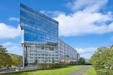 Totalbilde av stor kontorbygning med rektangelform og blank glassfasade, omgitt av grønn park og blå himmel i bakgrunnen.