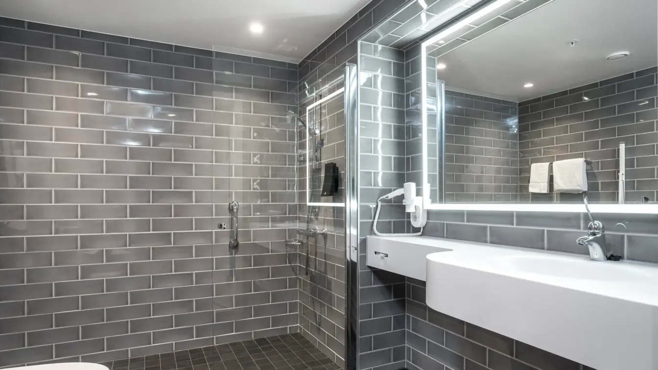 Vegger og gulv med grå fliser, dusjhjørne med glassvegg, speil med LED lys rundt og hvit servant. I superior baderom på Thon Hotel Høyers i Skien.