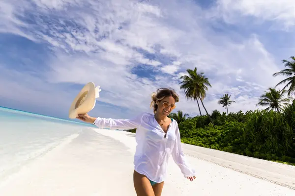 Halvportrett av smilende dame i bikini og hvit skjorte som går på eksotisk strand