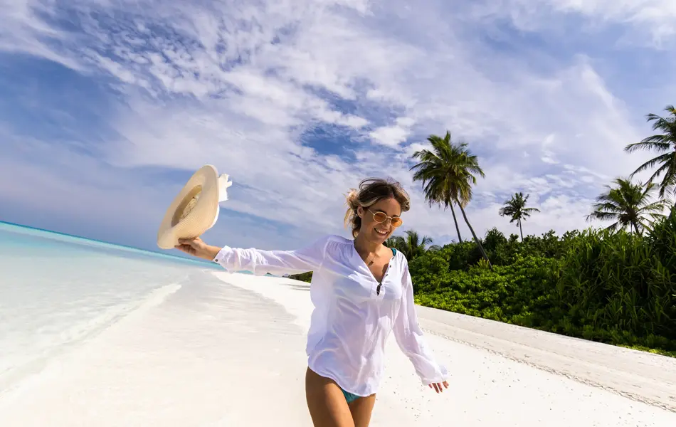Halvportrett av smilende dame i bikini og hvit skjorte som går på eksotisk strand