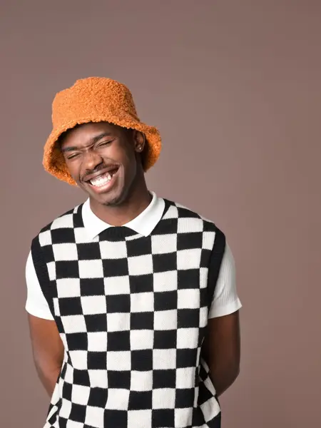 Mann i hvit og sortrutete vest, hvit t-skjorte og oransje teddybøttehatt smiler til kamera 
