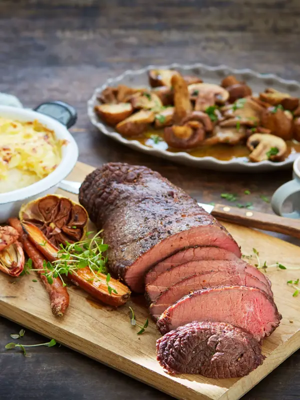 Fjøl med oppskåret hjortestek med fløtegratinerte poteter og tilbehør ved siden av