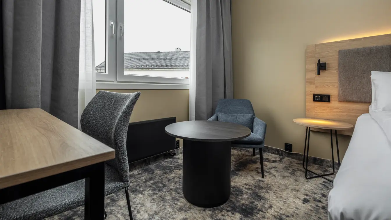 Dobbeltrom med sitteplass, pult og tilhørende stol, sengegavl i tre og grå gardiner.