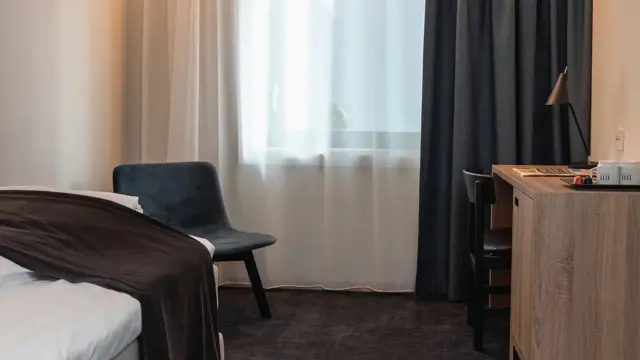 Mørkegrått gulvteppe, skrivebord i træ med sort stol, vindu med gardiner og en grå stol. I superior room på Thon Hotel Norge i Kristiansand.