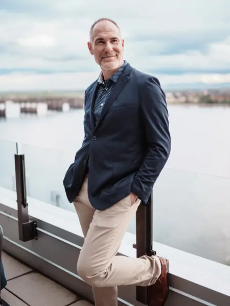 Mann med mørkeblå jakke og beige bukser lener seg inntil glassvegg foran sjø