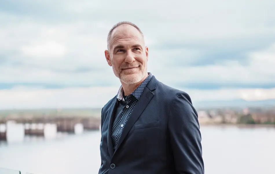 Mann med mørkeblå jakke og beige bukser lener seg inntil glassvegg foran sjø