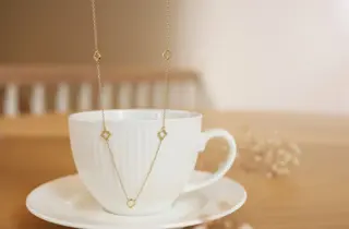 Et halssmykke med kløverdesign er holdt opp foran en kopp med kaffe.