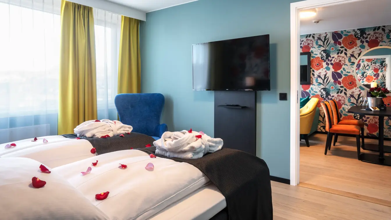 Vindu, dobbeltseng, morgenkåper, parkettgulv, TV, lyseblå vegger, blå stol, døråpning til stue, blomstertapetvegg, spisebord, oransje stoler