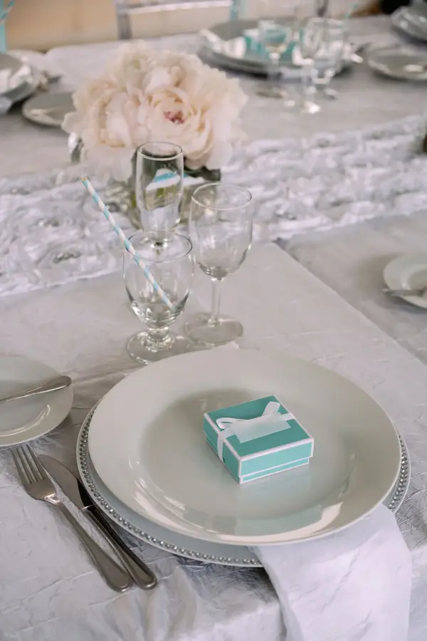 Dekorert bord med hvit duk, blondeløper, gule blomster og liten grønn boks på tallerkenen som navneskilt. Foto til artikkel om ting å huske på når du skal arrangere konfirmasjon.