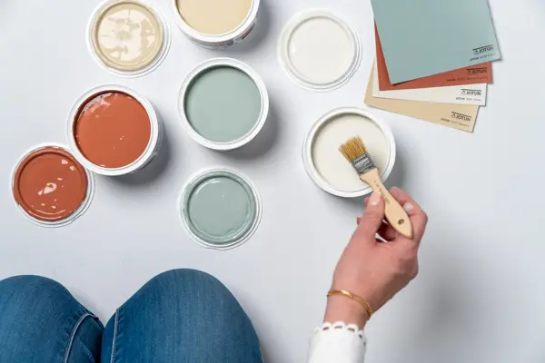 En dame som holder på med oppussing og tips om interiør trender for våren 2021. Damen har lyst halvlangt hå og har på seg en blå olaskjorte og blå olabukse. Fargene går i brunt og beige jordtoner og det er mye lilla og pastellfarger. Vårens interiør