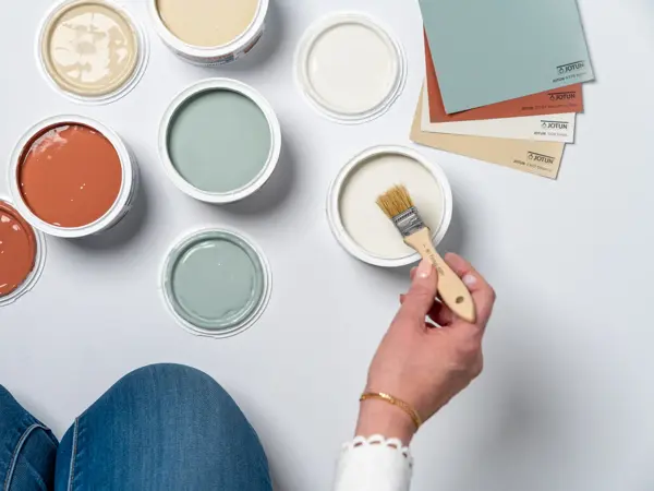 En dame som holder på med oppussing og tips om interiør trender for våren 2021. Damen har lyst halvlangt hå og har på seg en blå olaskjorte og blå olabukse. Fargene går i brunt og beige jordtoner og det er mye lilla og pastellfarger. Vårens interiør