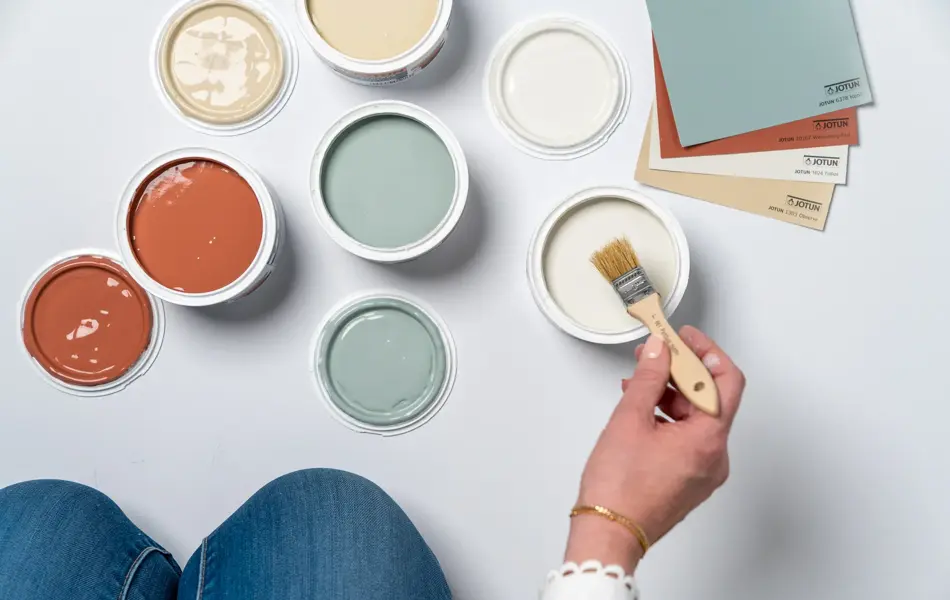 En dame som holder på med oppussing og tips om interiør trender for våren 2021. Damen har lyst halvlangt hå og har på seg en blå olaskjorte og blå olabukse. Fargene går i brunt og beige jordtoner og det er mye lilla og pastellfarger. Vårens interiør