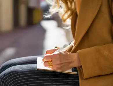 Jente som sitter ute i kamelfarget jakke og stripete bukser og skriver