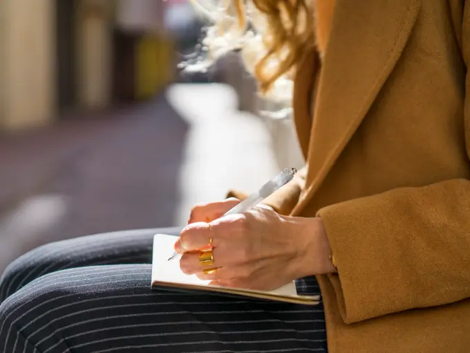 Jente som sitter ute i kamelfarget jakke og stripete bukser og skriver