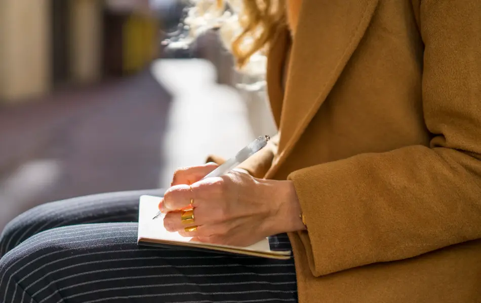 Jente som sitter ute i kamelfarget jakke og stripete bukser og skriver