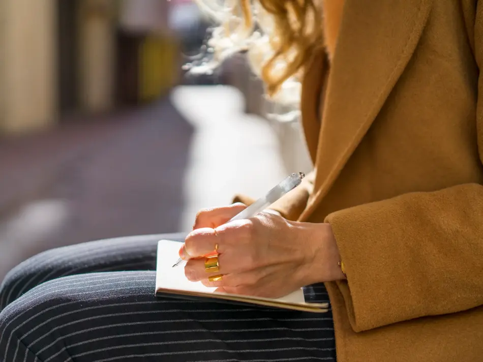 Jente som sitter ute i kamelfarget jakke og stripete bukser og skriver