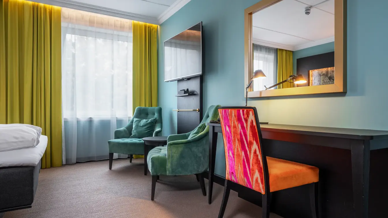 Businessrom med dobbeltseng, skrivebord, oransje stol, speil, vegghengt TV, gule gardiner og to grønne lenestoler