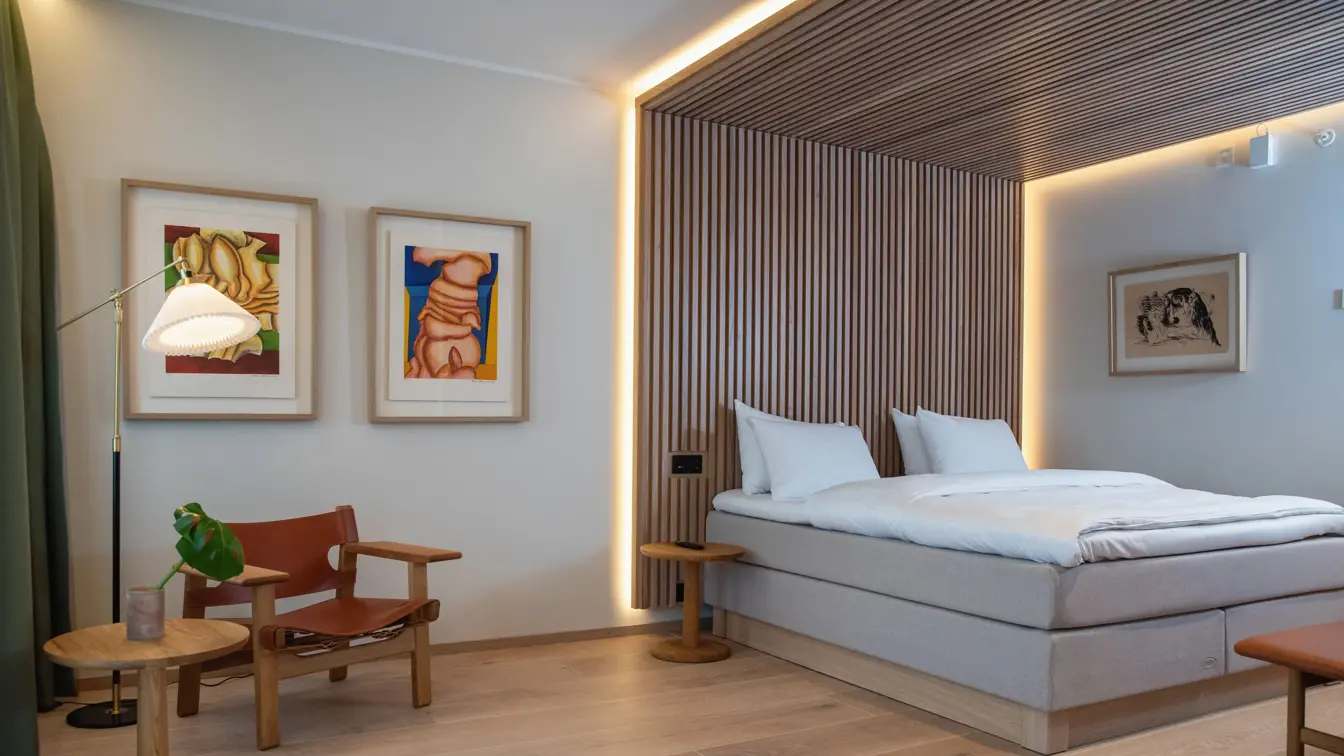 Dobbeltseng med stripete tapet som sengegavl og i tak over sengen. To fargerike bilder på hvit vegg. En stol i træ og skinn og lite rundt bord.  I suite på Thon Hotel Norge i Kristiansand.