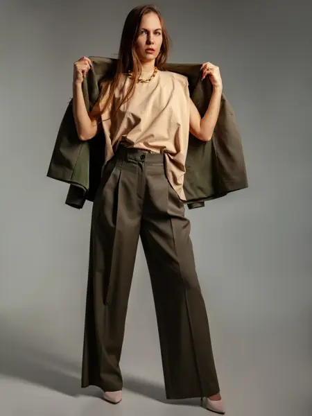 En kvinne poserer i et fotostudio. Hun er iført en brun dressbukse med høyt liv, høyhælte sko og en beige topp. Hun holder dressjakken over skuldrene. Håret er brunt og henger løst nedover skuldrene.