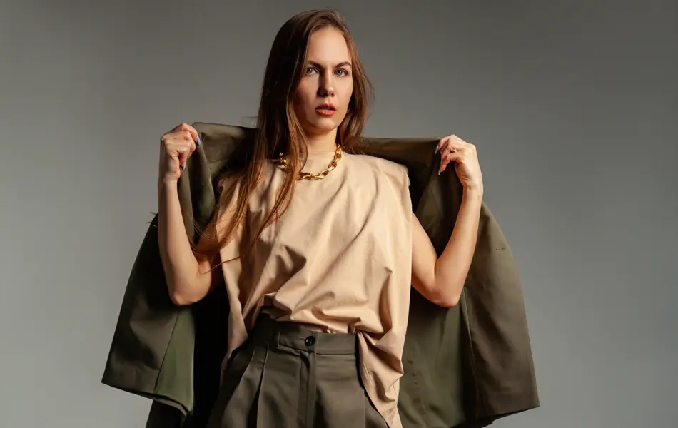 En kvinne poserer i et fotostudio. Hun er iført en brun dressbukse med høyt liv, høyhælte sko og en beige topp. Hun holder dressjakken over skuldrene. Håret er brunt og henger løst nedover skuldrene.