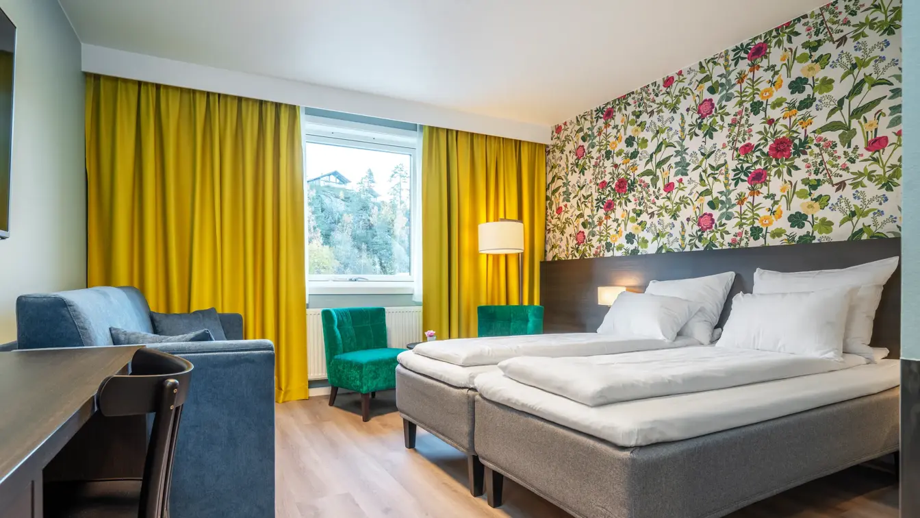 Familierom med twin senger, blå sofa, skrivebord med stol, blomstrete tapet, grønne lenestoler og gule gardiner.