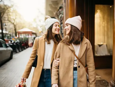 To jenter i beige kåpe smiler mot hverandre på gata