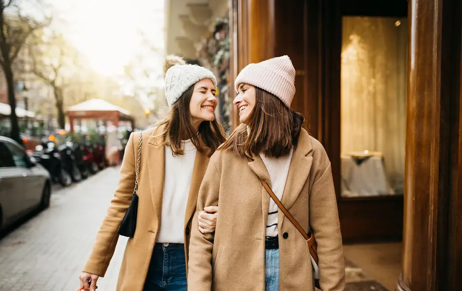 To jenter i beige kåpe smiler mot hverandre på gata