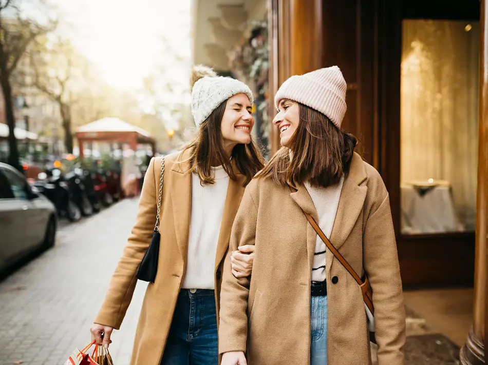 To jenter i beige kåpe smiler mot hverandre på gata