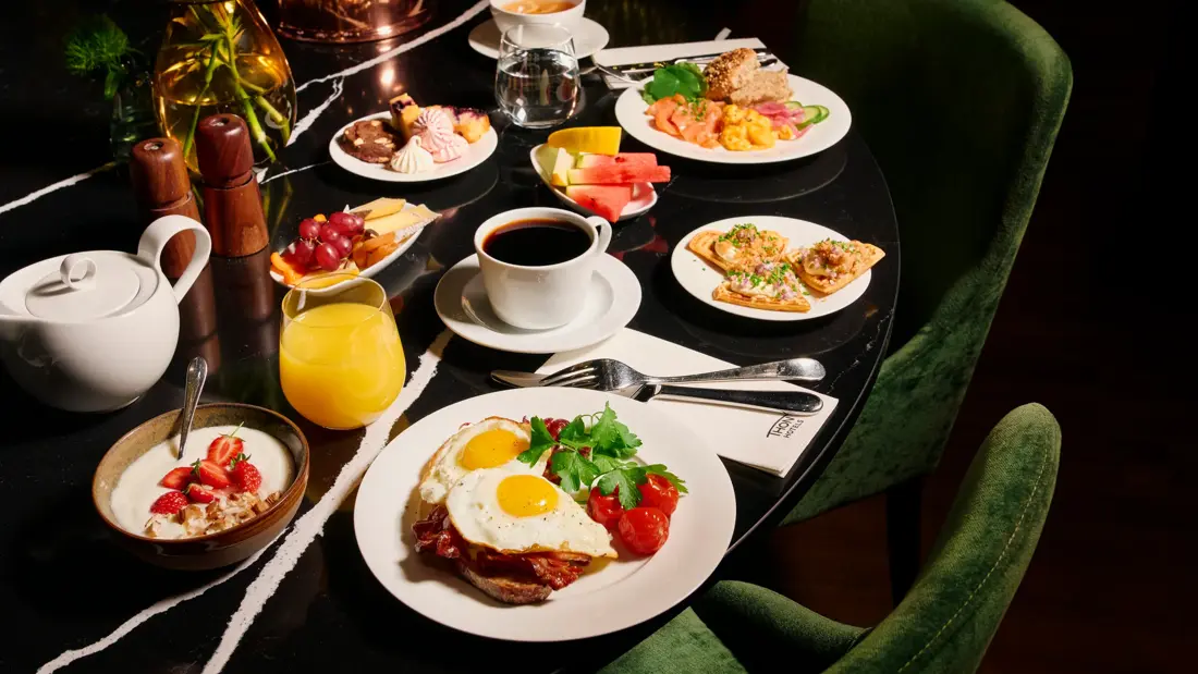 Et bugnende frokostbord med ulike retter, inkludert speilegg med bacon, røkelaks med eggerøre, vafler med rogn, kaker, yoghurt med jordbær, oster og frukt. Drikkevarer inkluderer kaffe, te, vann og juice. Mørkegrønne stoler rundt bordet.