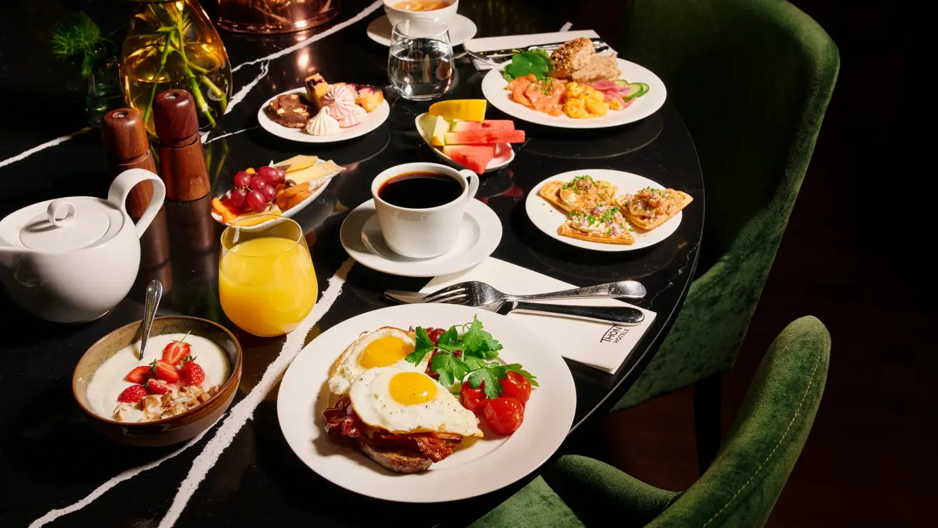 Et bugnende frokostbord med ulike retter, inkludert speilegg med bacon, røkelaks med eggerøre, vafler med rogn, kaker, yoghurt med jordbær, oster og frukt. Drikkevarer inkluderer kaffe, te, vann og juice. Mørkegrønne stoler rundt bordet.