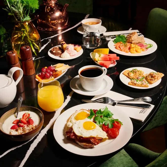 Et bugnende frokostbord med ulike retter, inkludert speilegg med bacon, røkelaks med eggerøre, vafler med rogn, kaker, yoghurt med jordbær, oster og frukt. Drikkevarer inkluderer kaffe, te, vann og juice. Mørkegrønne stoler rundt bordet.