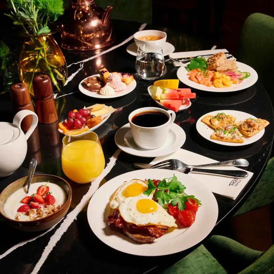 Et bugnende frokostbord med ulike retter, inkludert speilegg med bacon, røkelaks med eggerøre, vafler med rogn, kaker, yoghurt med jordbær, oster og frukt. Drikkevarer inkluderer kaffe, te, vann og juice. Mørkegrønne stoler rundt bordet.