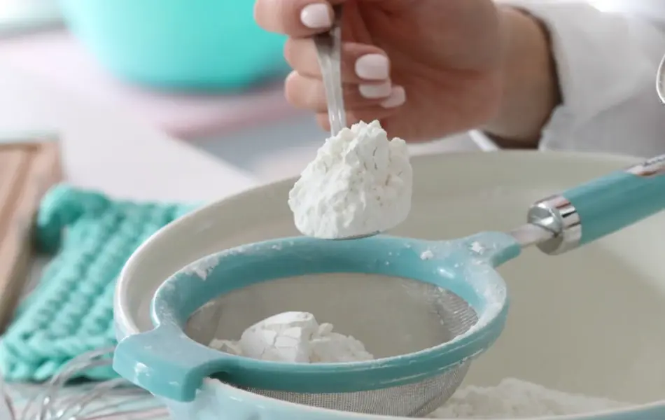 Hånd som holder en skje med mel over en bakebolle med sil