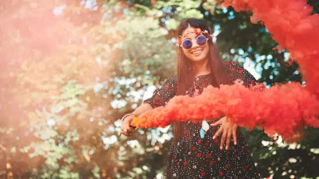 Jente med blomsterkrans og runde solbriller holder en rød/oransje fargebombe