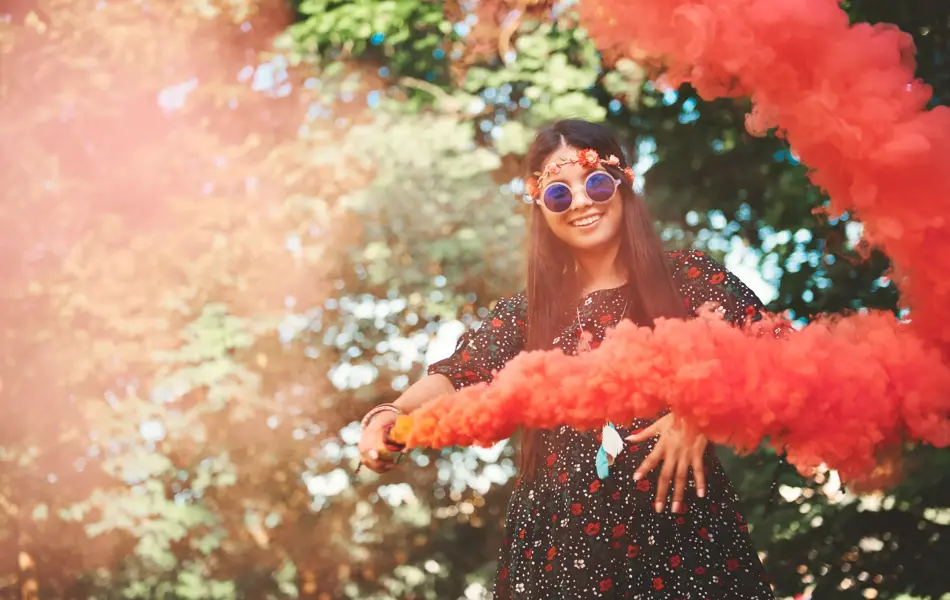 Jente med blomsterkrans og runde solbriller holder en rød/oransje fargebombe