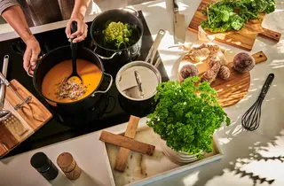 En person står ved en ovn og lager mat i tre kjeler, mens det er flere ingredienser liggende på skjærefjøler rundt på kjøkkenbenken.