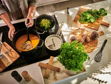 En person står ved en ovn og lager mat i tre kjeler, mens det er flere ingredienser liggende på skjærefjøler rundt på kjøkkenbenken.
