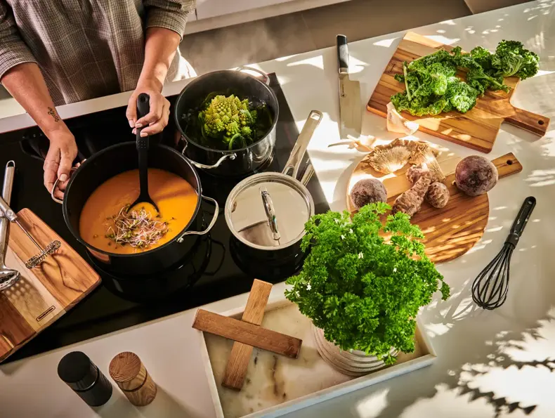 En person står ved en ovn og lager mat i tre kjeler, mens det er flere ingredienser liggende på skjærefjøler rundt på kjøkkenbenken.