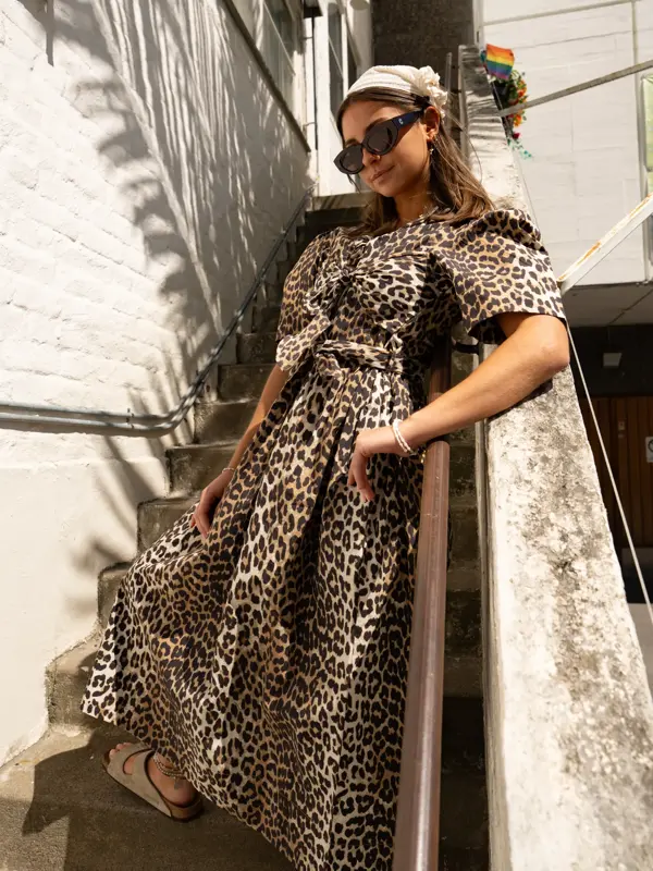 Jente står utendørs i trapp med hvitt hårtørkle, leopardmønstret lang kjole og semsket beige Birckenstock.