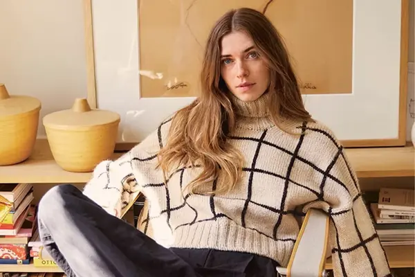 dame med lang brunt hår sitter i stol, hvit genser med svarte striper, mørk bukse