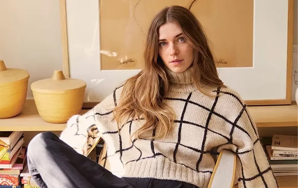 dame med lang brunt hår sitter i stol, hvit genser med svarte striper, mørk bukse