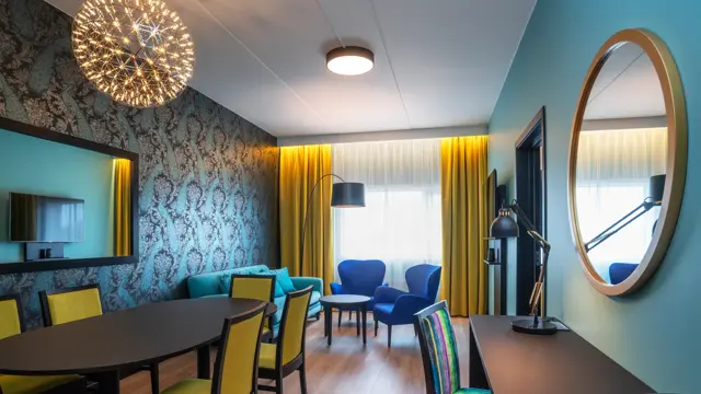 Tapetvegger med mønster, speil på veggen, vindu fra gulv til tak, gul gardin, turkis sofa, to blå stoler, parkettgulv, langt spisebord med gule stoler. Svart skrivebord med stol.