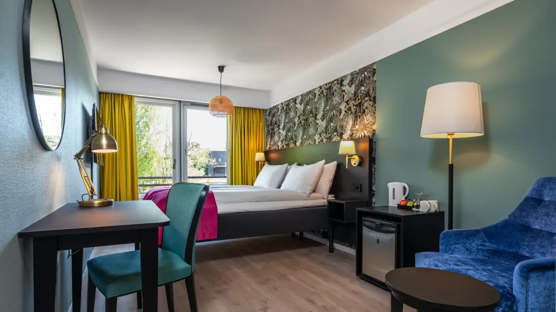 Standard rom med twin senger, rosa sengeteppe, skrivebord med tilhørende stol, gule gardiner, stort vindu og minibar.