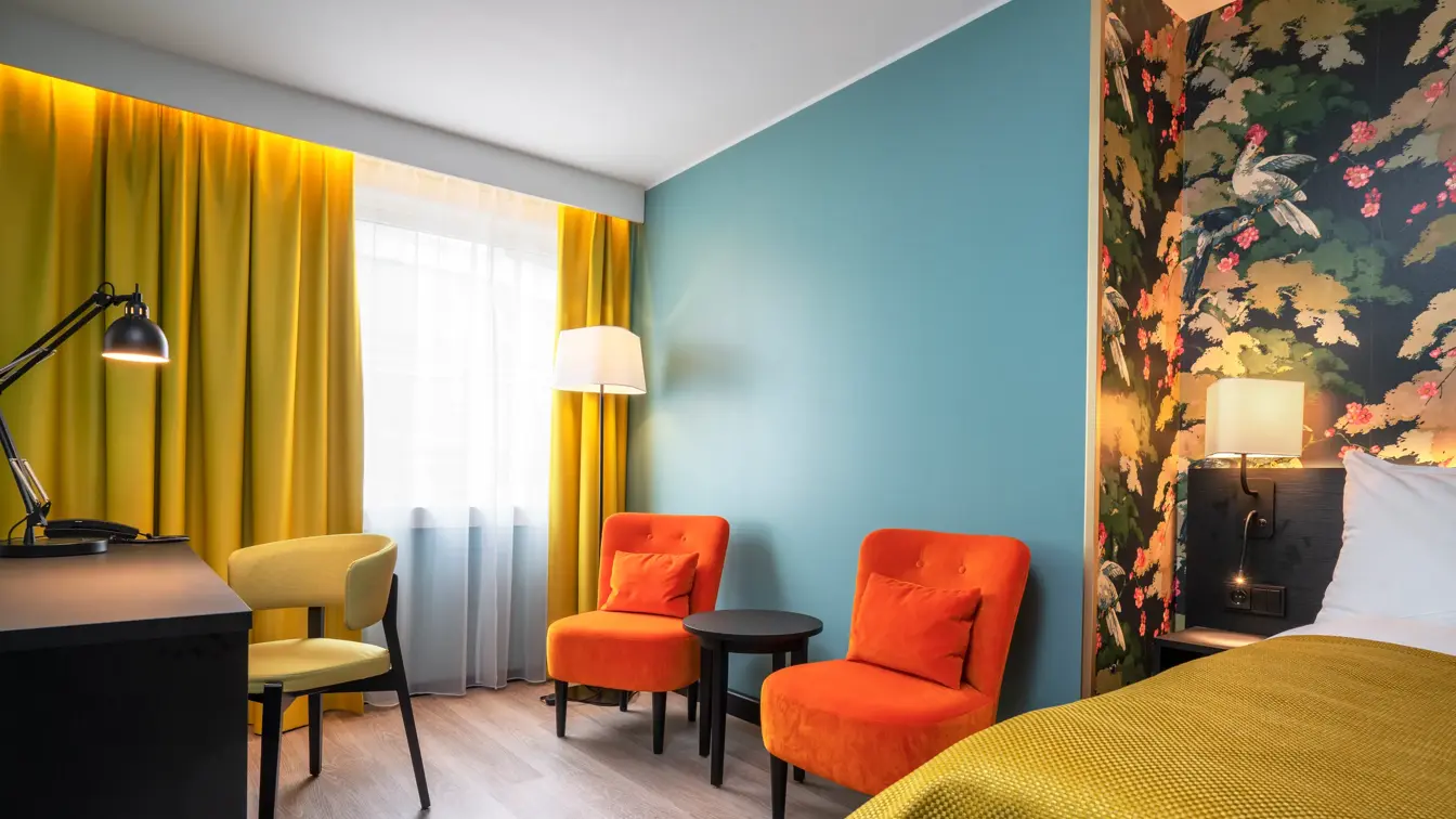 Fargerik tapet og dobbeltseng med gult sengeteppe. Turkis tapet, oransje stoler, mørkt rundt bord og gule gardiner. Skrivebord med leselampe og gul stol i dobbeltrom på Thon Hotel Bristol Bergen